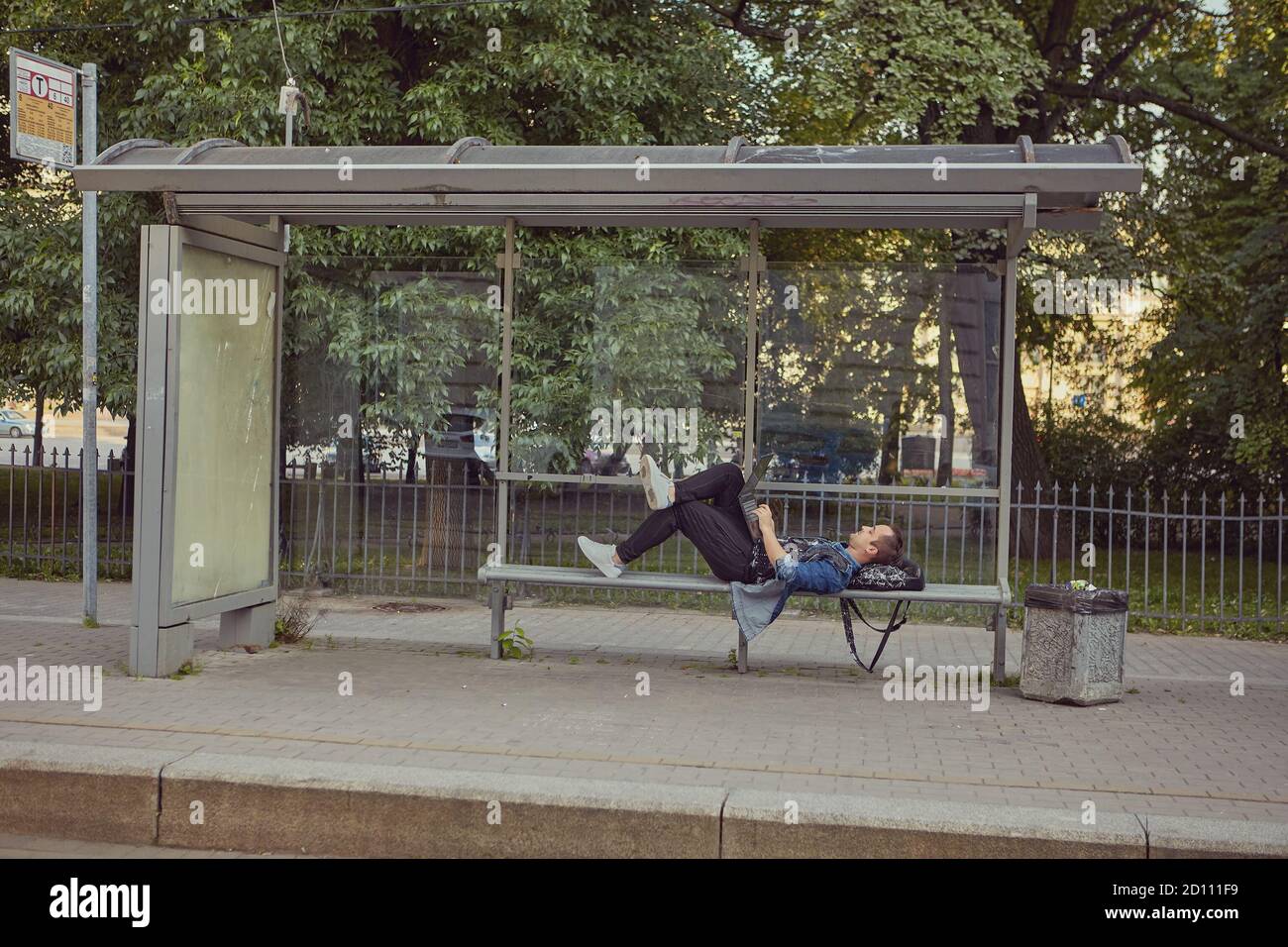 Un homme blanc à l'arrêt de bus s'est posé sur un banc avec un ordinateur portable à genoux. Un jeune homme utilise un ordinateur portable tout en attendant les transports en commun. Banque D'Images