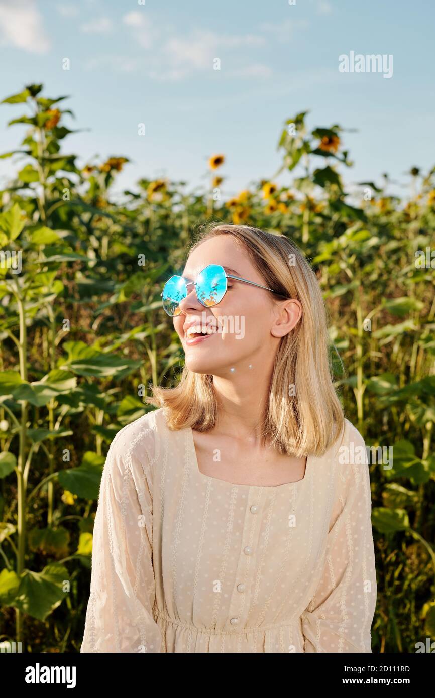 Joyeuse jeune femme blonde en lunettes de soleil et robe blanche debout dans le champ Banque D'Images