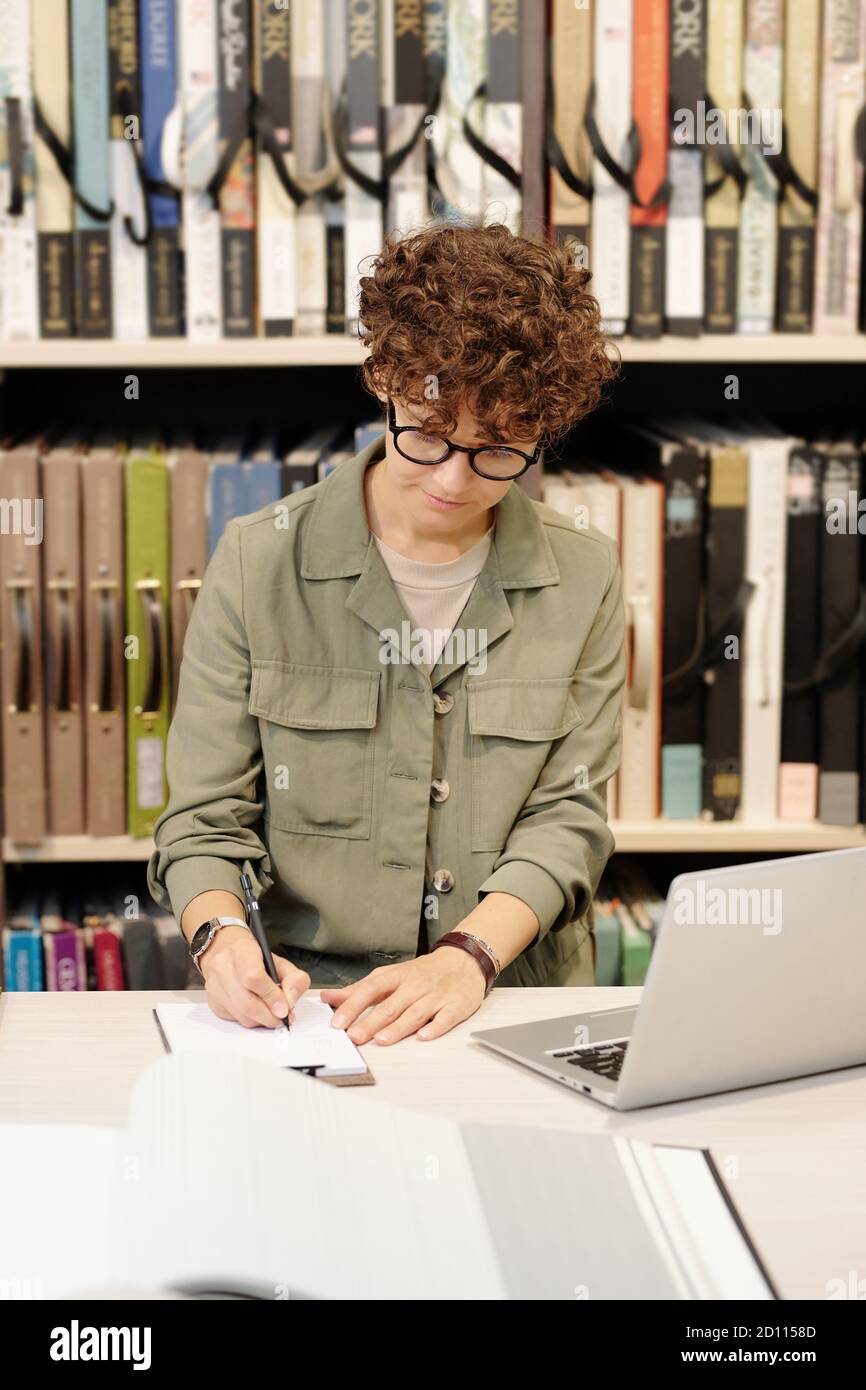 Jeune assistant de magasin contemporain qui prend des notes dans un ordinateur portable à l'avant de l'ordinateur portable Banque D'Images