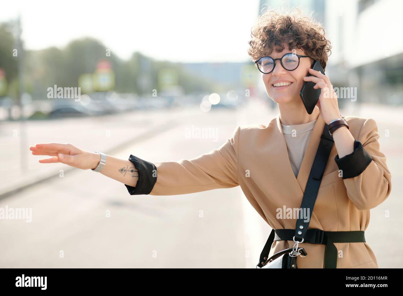 Bonne jeune femme élégante avec smartphone à l'oreille prenant le taxi par la route Banque D'Images