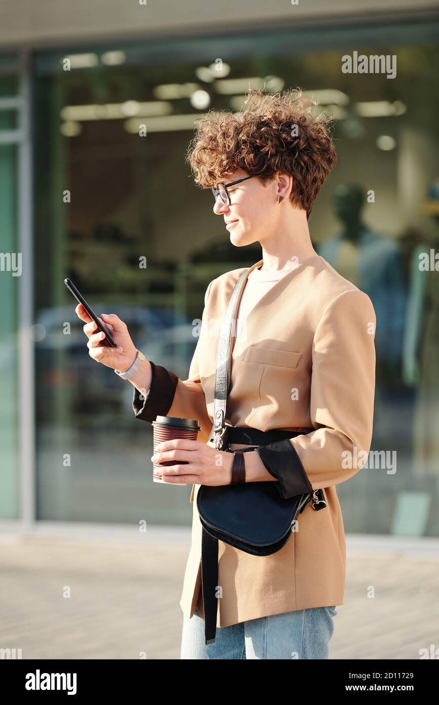Jeune femme d'affaires brunette avec boisson et sac à main textant en mobile téléphone Banque D'Images