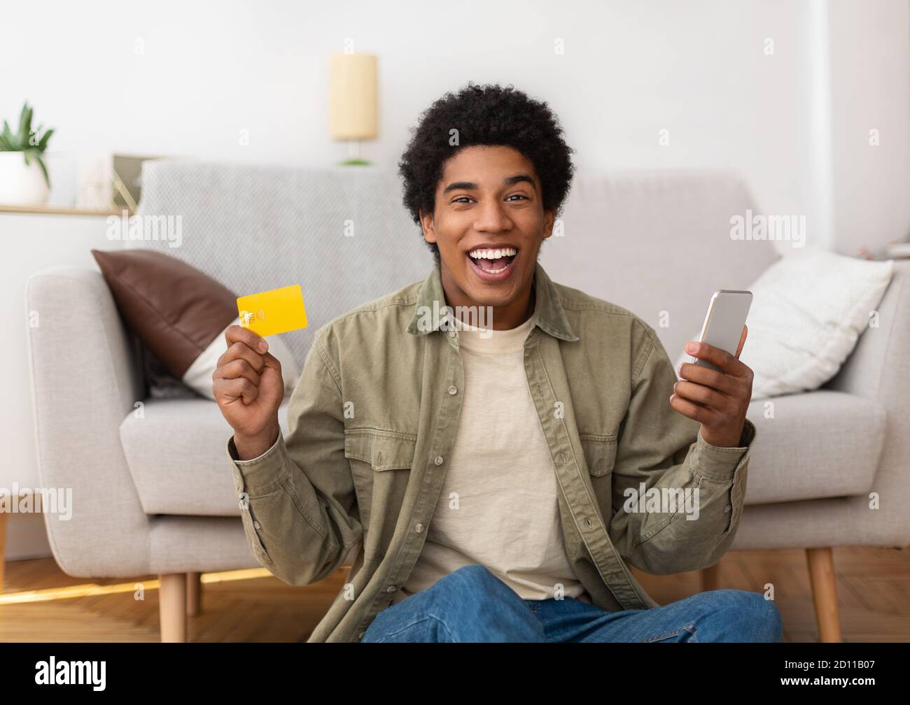 Cybershopping. Un adolescent afro-américain enthousiaste avec téléphone portable et carte de crédit commandant des produits en ligne à partir de la maison Banque D'Images