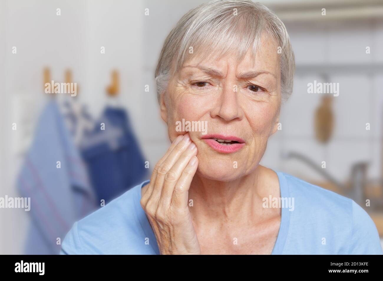 Mal de dents aigu : femme âgée dans sa cuisine avec une main à sa mâchoire douloureuse. Banque D'Images