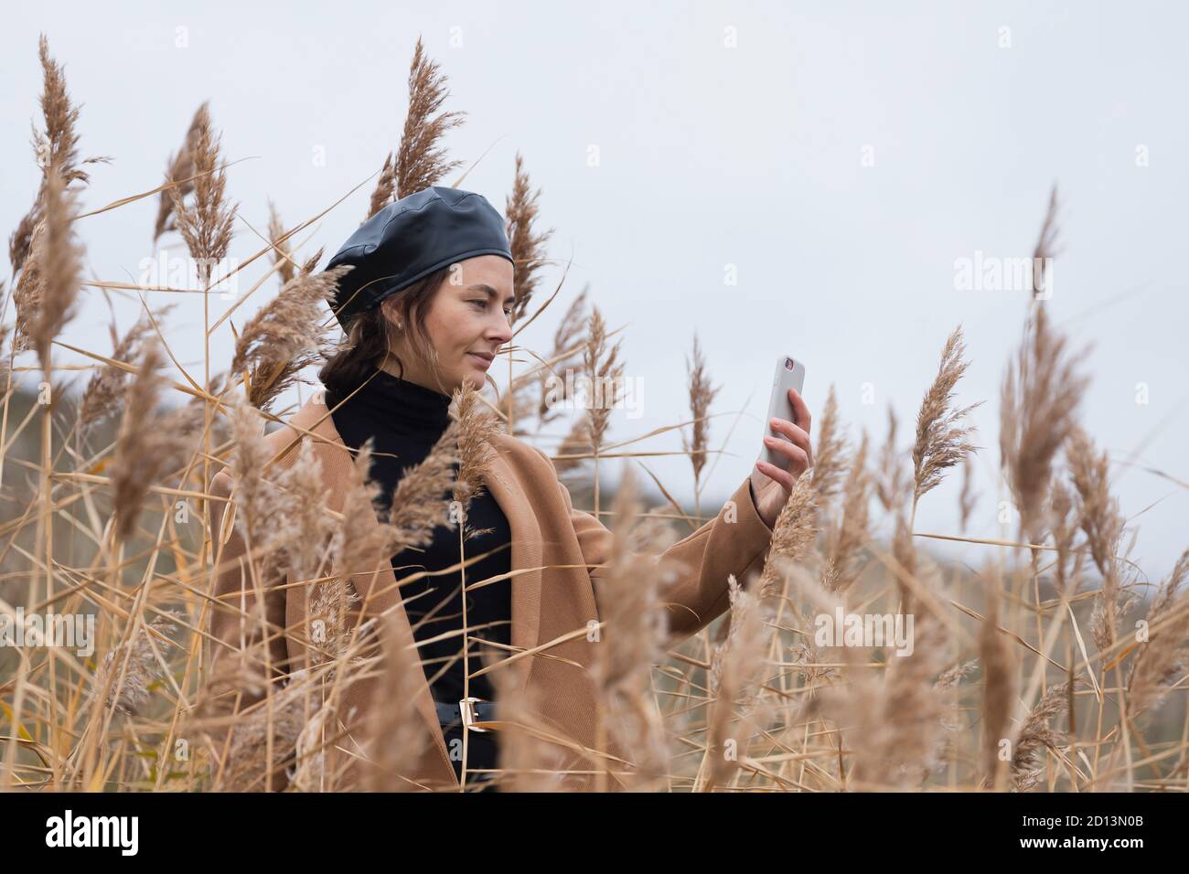 Une femme gaie aux cheveux sombres dans un béret noir brillant et un manteau de laine beige souriant, prend un selfie profiter de la journée, sur le fond dans le soleil de l'automne da Banque D'Images