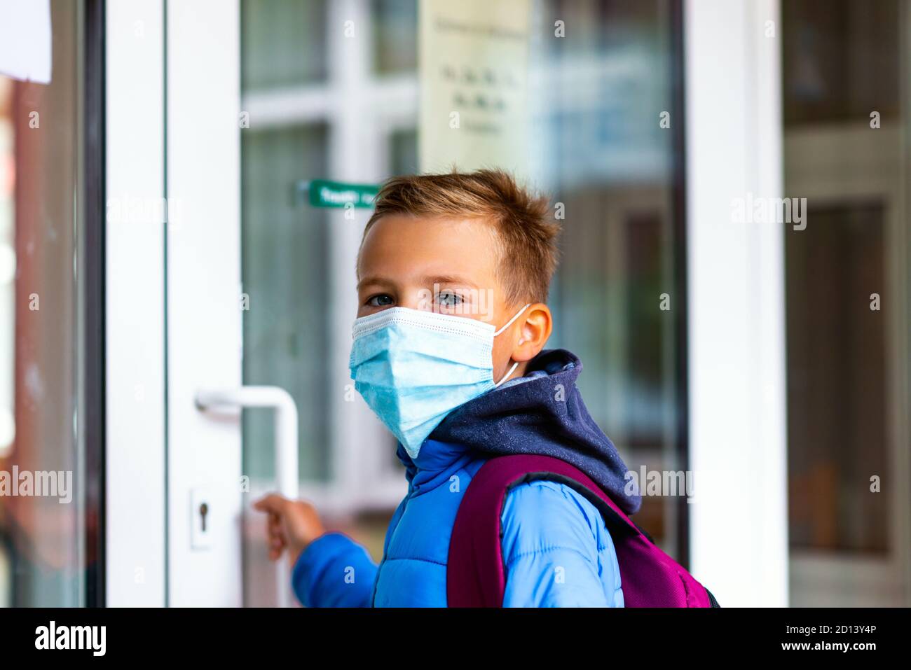 Un jeune écolier portant un masque de protection tente d'ouvrir la porte de l'école. Derrière le sac à dos, l'écolier regarde l'appareil photo Banque D'Images