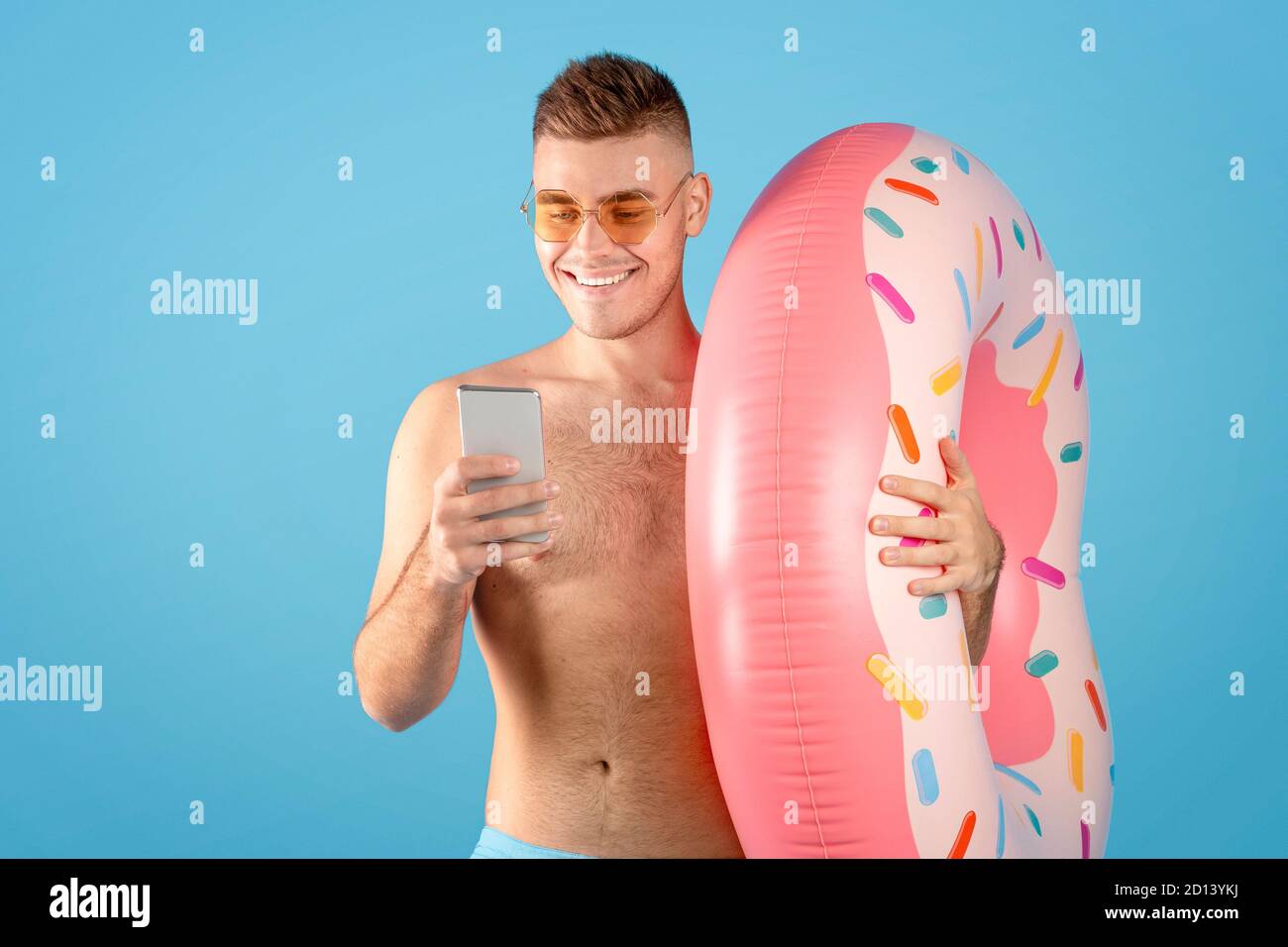 Agence de tourisme en ligne. Jeune homme avec anneau gonflable et smartphone réservant visite tropicale sur le web, fond bleu Banque D'Images