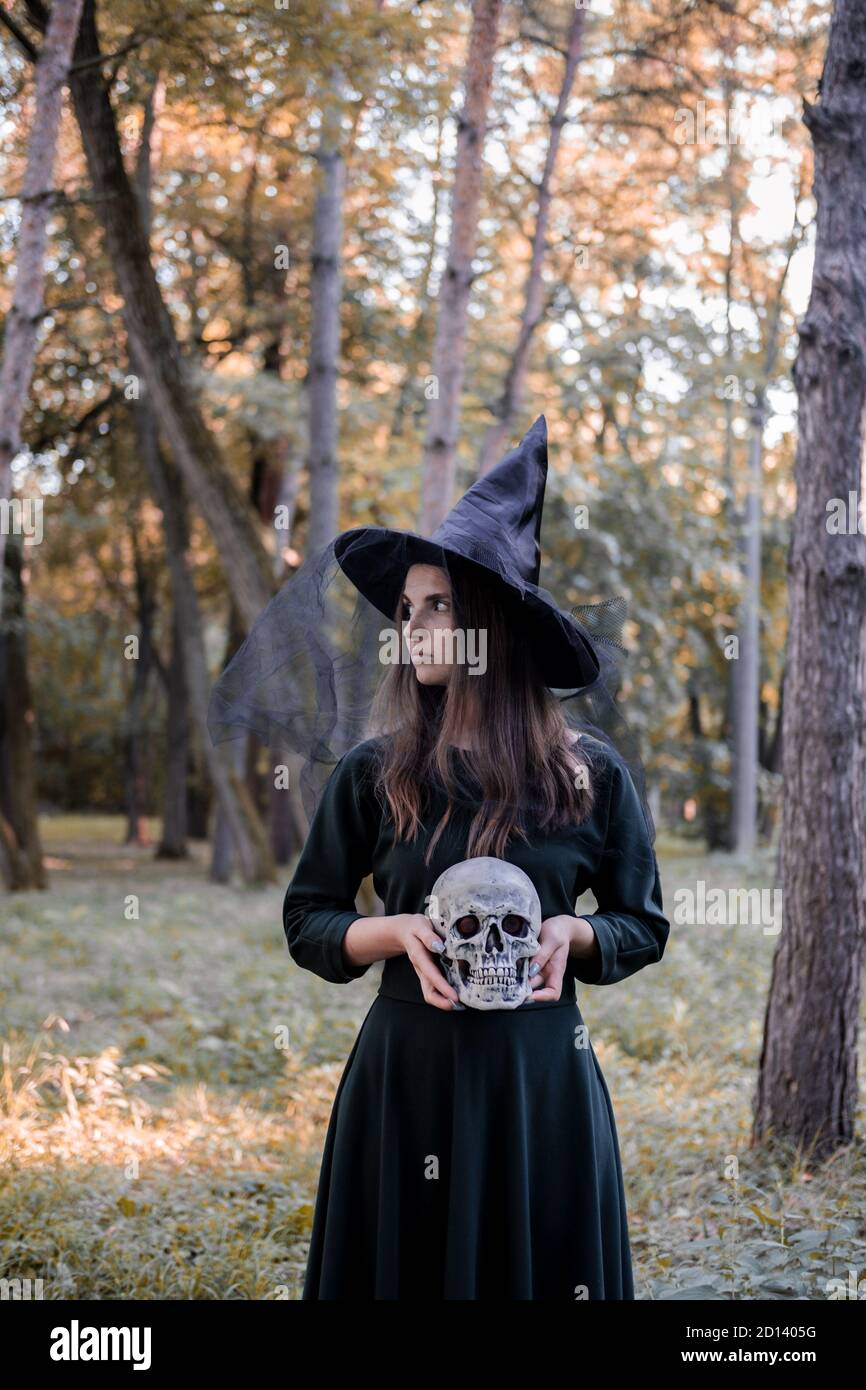 Jeune belle femme mignonne en robe sombre et chapeau de sorcière tient un crâne dans ses mains. Costume de fête d'Halloween. Forêt, parc avec arbres d'automne Banque D'Images