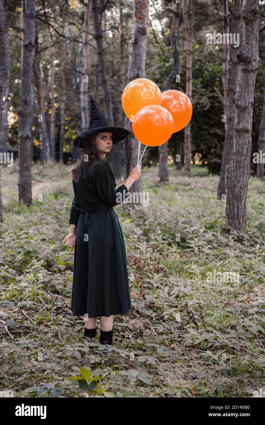 Jeune mignonne belle femme en robe sombre et chapeau de sorcière tient des ballons d'orange dans ses mains. Costume de fête d'Halloween. Forêt, parc avec arbres d'automne Banque D'Images