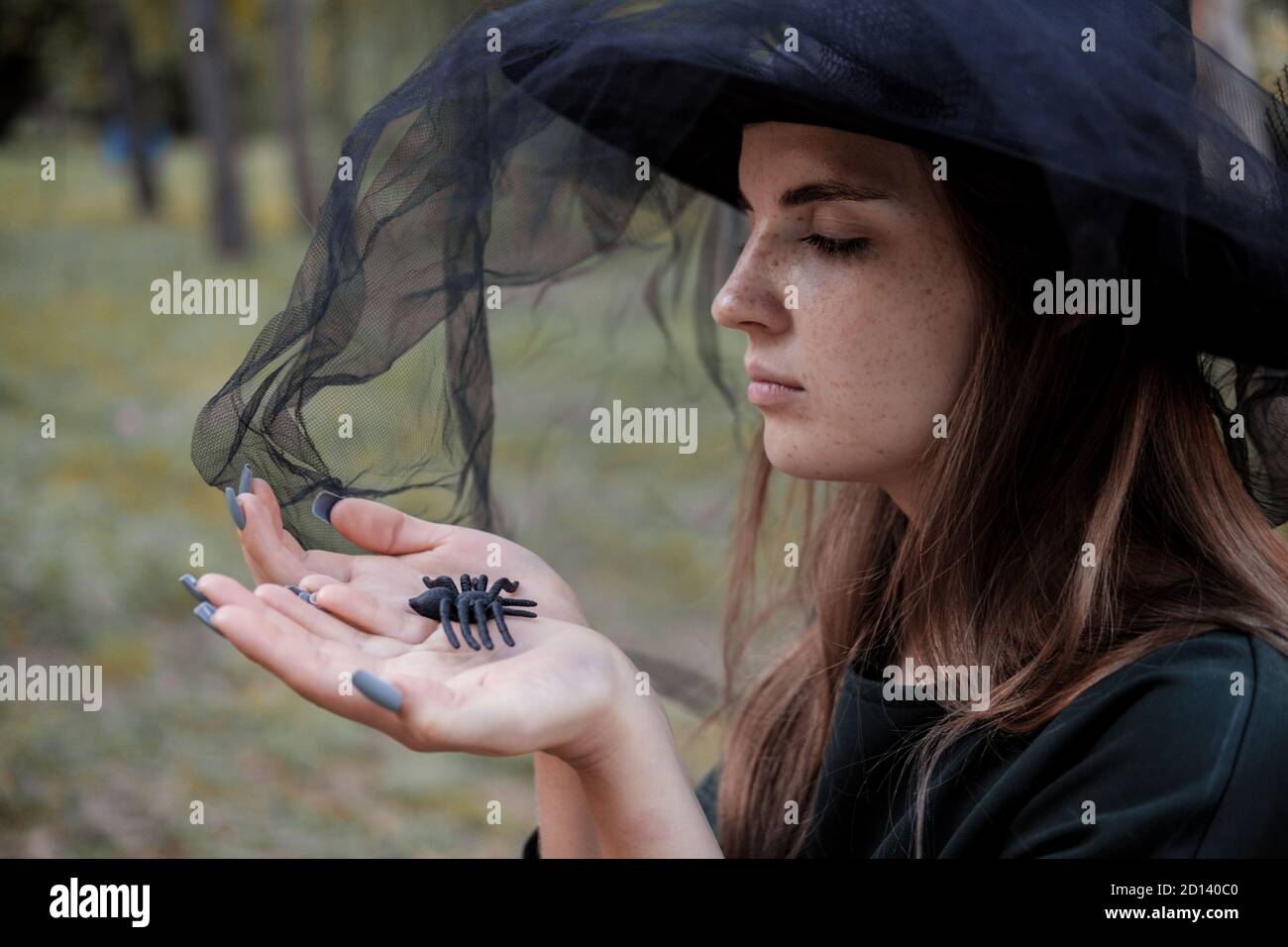 Jeune mignonne belle femme en robe sombre et chapeau de sorcière hotsblack araignée dans ses mains. Costume de fête d'Halloween. Forêt, parc avec arbres d'automne Banque D'Images