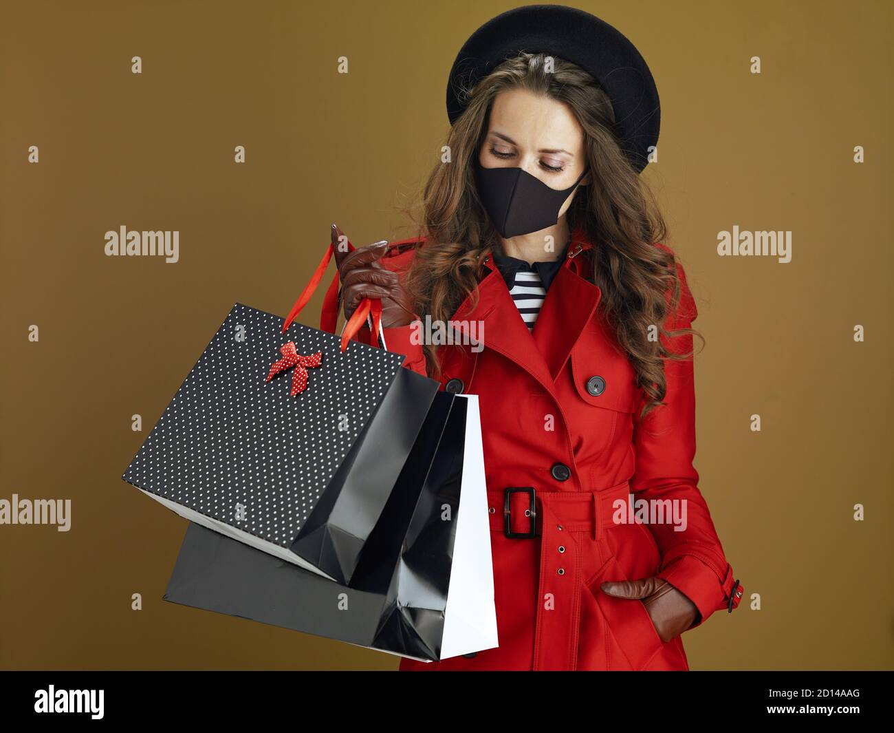 La vie durant la pandémie du coronavirus. Femme tendance de 40 ans en manteau rouge avec masque noir et sacs de shopping en papier sur fond de bronze. Banque D'Images