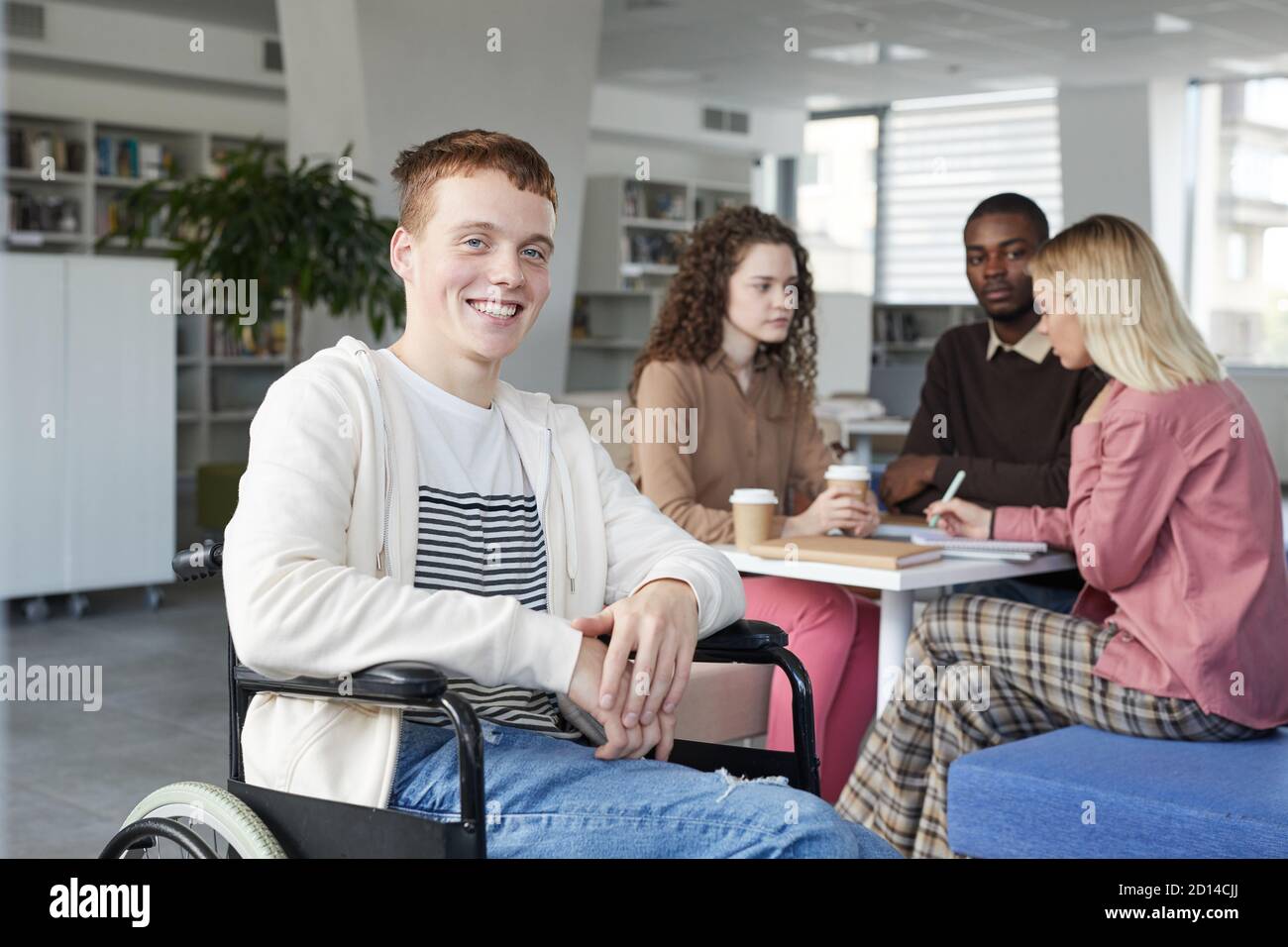 Portrait d'un garçon souriant aux cheveux rouges en fauteuil roulant étudiant avec un groupe d'étudiants dans la bibliothèque de l'université et regardant l'appareil photo, l'espace de copie Banque D'Images