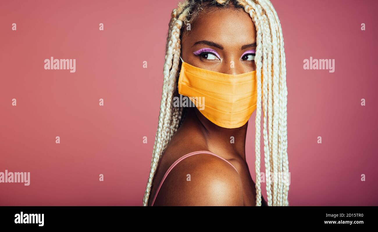 Jeune femme africaine avec des tresses blondes en boîte portant un masque facial de protection. Femme avec style africain de cheveux et masque de visage regardant loin sur fond rose. Banque D'Images