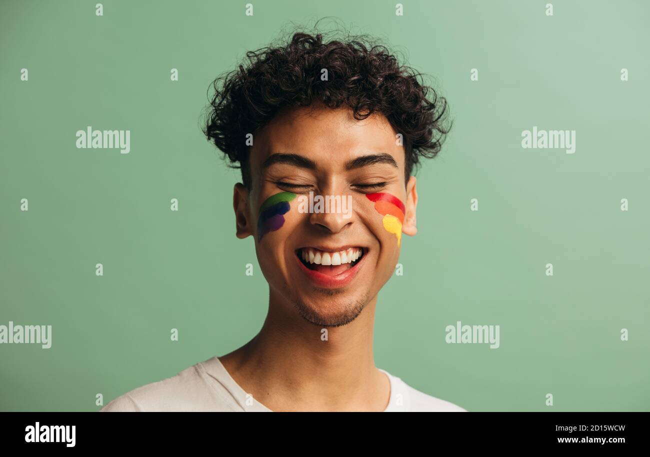 Gros plan d'un jeune homme joyeux avec un drapeau de fierté peint sur le visage. Homme gay avec visage arc-en-ciel peint sourire sur fond vert pastel. Banque D'Images