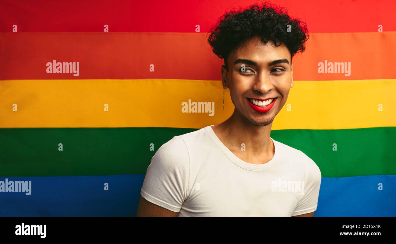Portrait d'un bel homme gay portant le maquillage contre le drapeau de fierté. Homme liquide de sexe avec lèvre rouge et contour d'oreille regardant loin et souriant. Banque D'Images