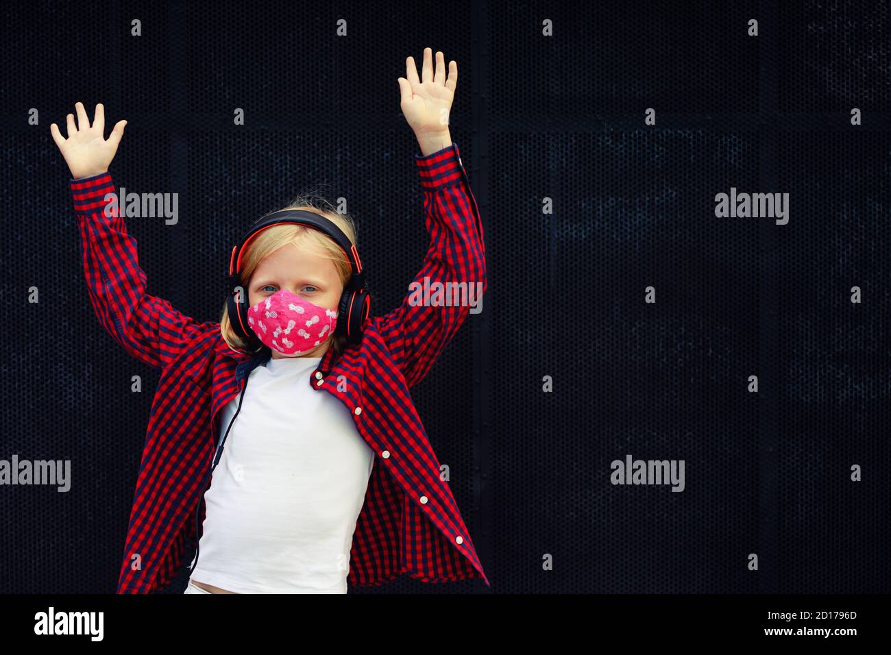 Un jeune enfant positif dans un casque qui écoute de la musique avec plaisir lève les mains. Drôle fille porter élégant masque de protection du visage due coronavirus COVID 19 Banque D'Images