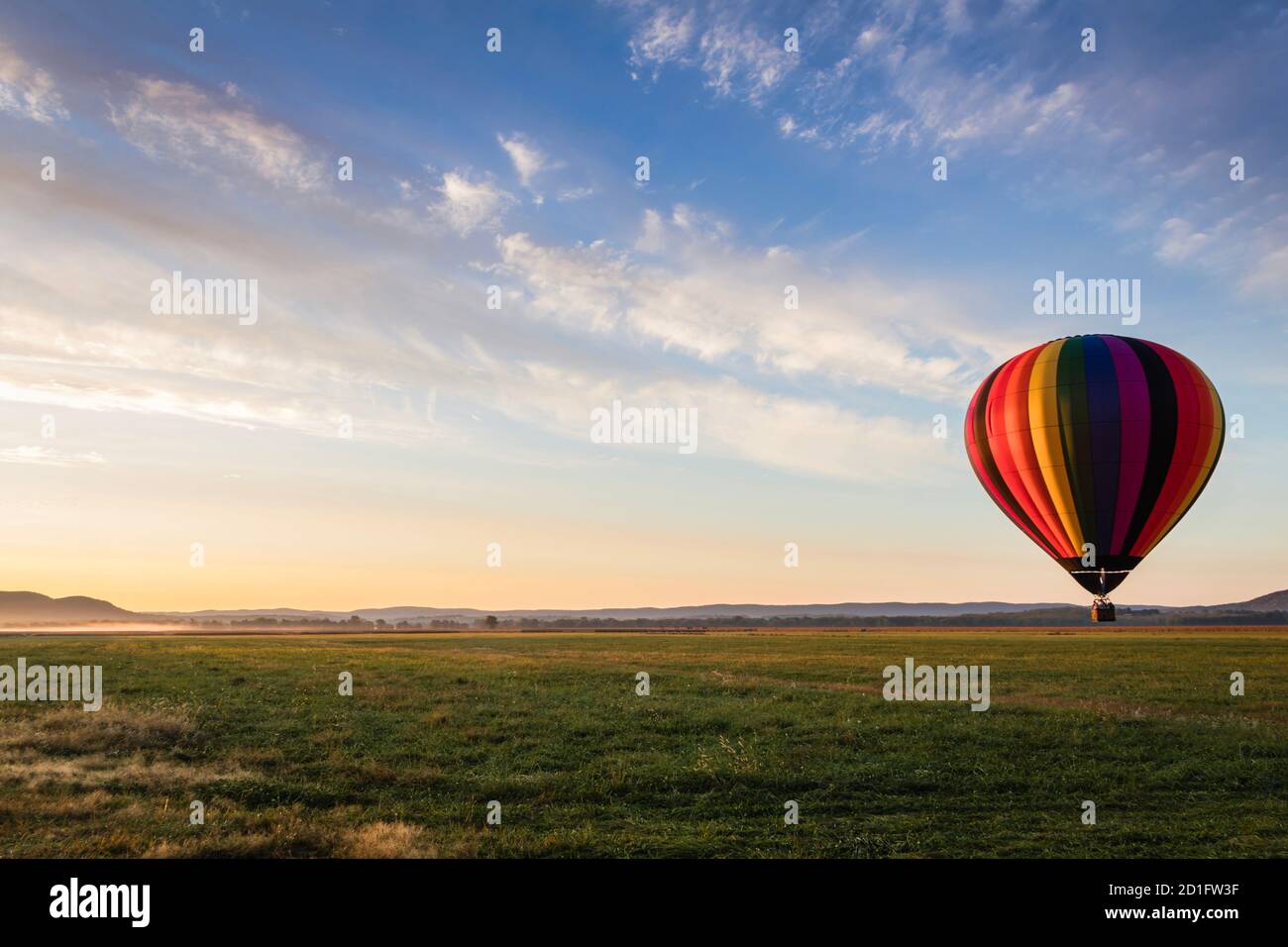 Le ballon d'air chaud dans des bandes arc-en-ciel colorées commence l'ascension champ de ferme au soleil se lève ciel bleu ciel nuageux Banque D'Images
