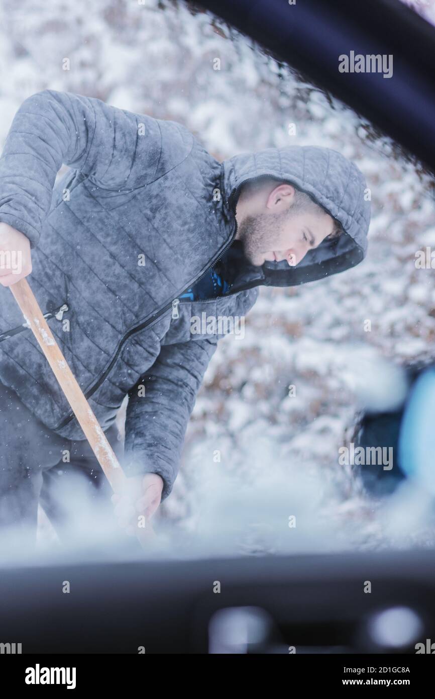 Vue de l'intérieur de la voiture sur un homme vêtu d'une veste qui nettoie la neige autour de sa voiture. Banque D'Images