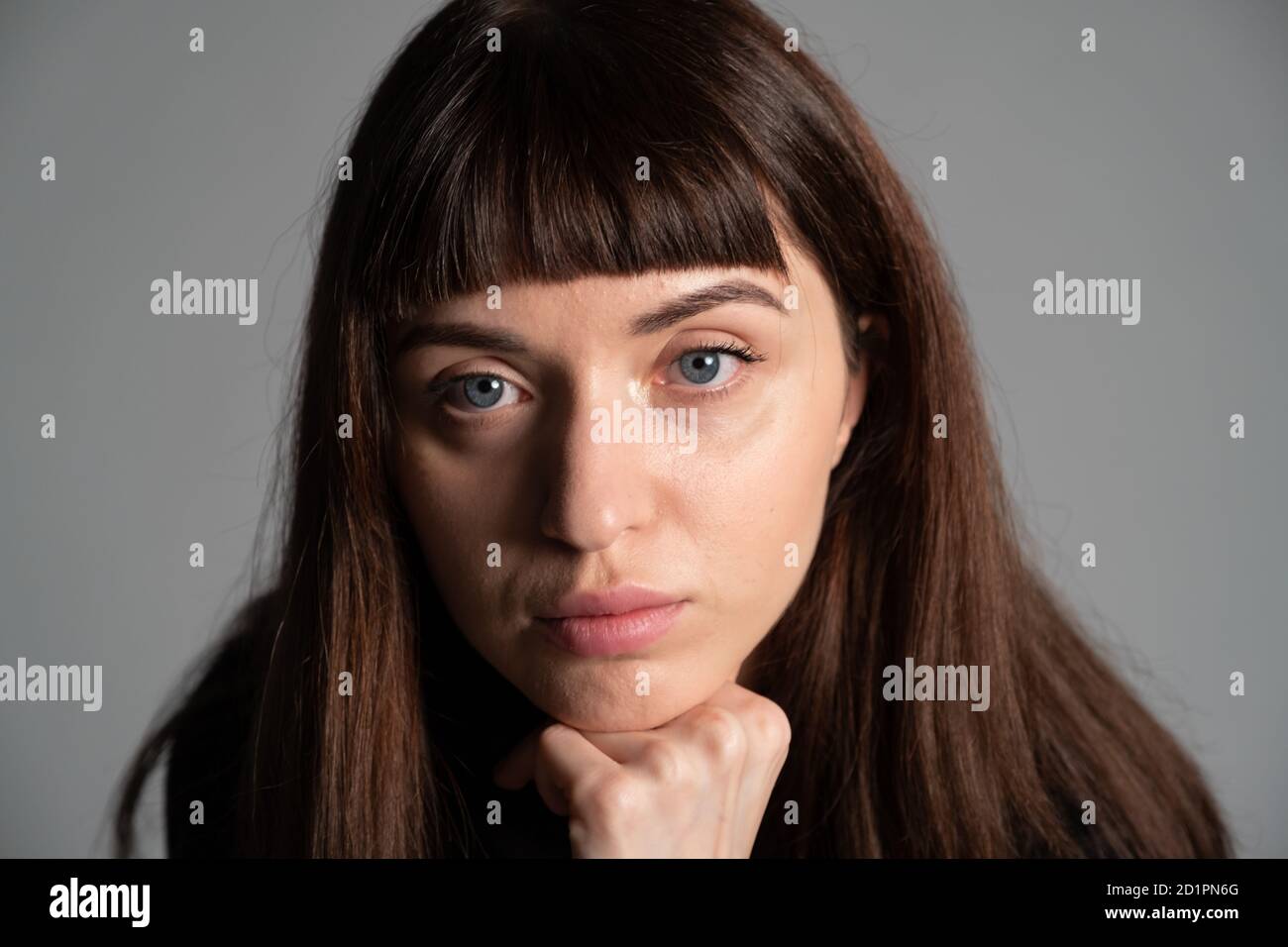 Gros plan portrait en studio d'une jolie femme brune, main sur le menton, regardant sérieusement l'appareil photo, sur un fond gris Uni Banque D'Images
