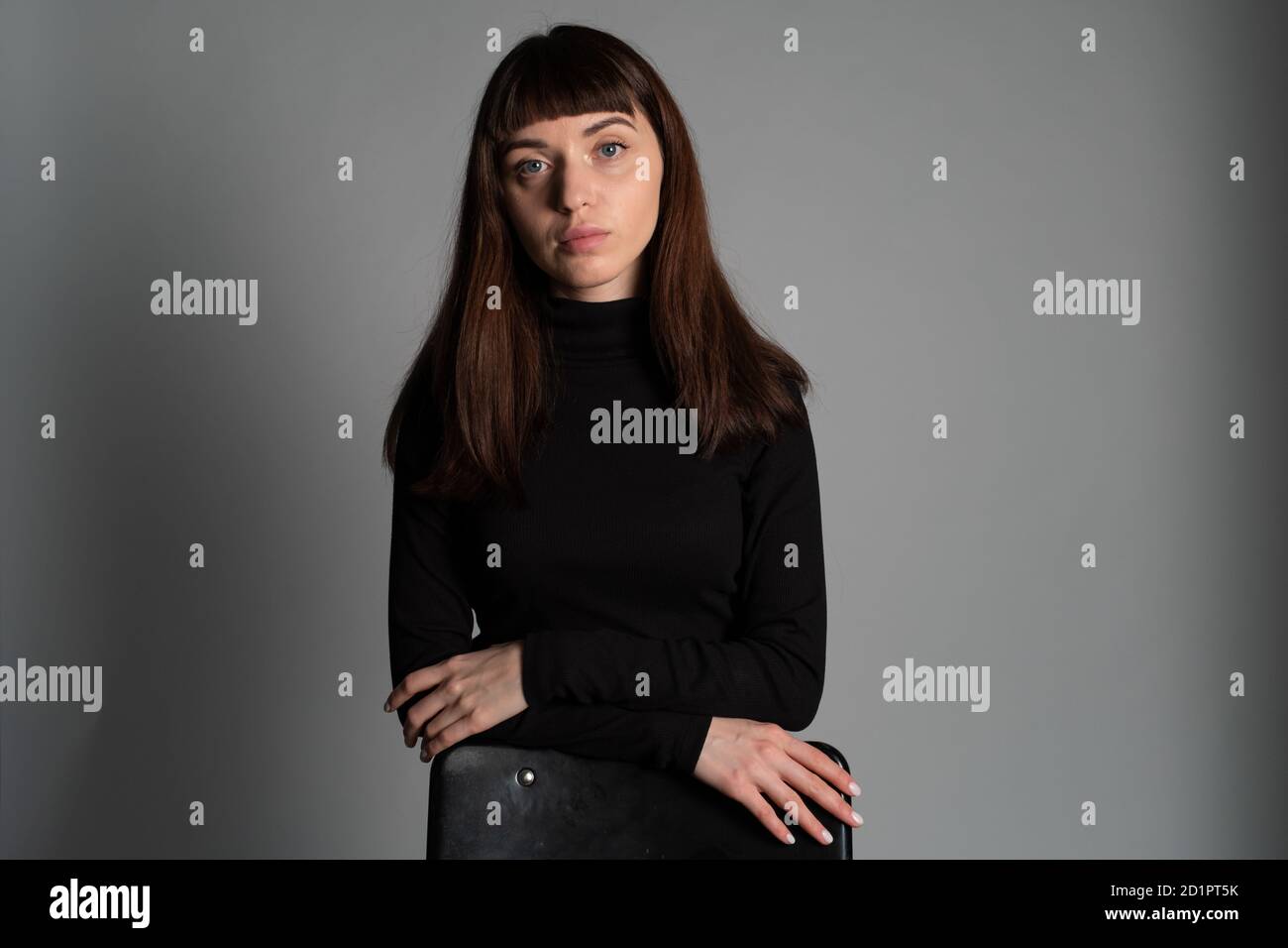 Gros plan studio portrait d'une jolie femme brune, debout derrière la chaise avec des bras croisés, regardant sérieusement la caméra, contre un GRE simple Banque D'Images