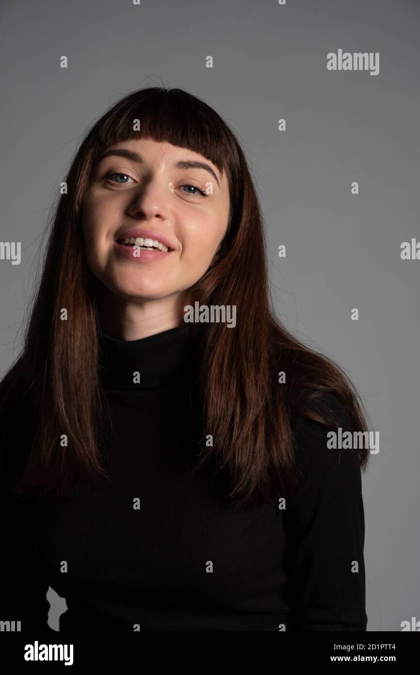 Gros plan portrait en studio d'une jolie femme brune, portant un pull à col roulé noir plié, souriant, regardant l'appareil photo, contre un fond gris Uni Banque D'Images