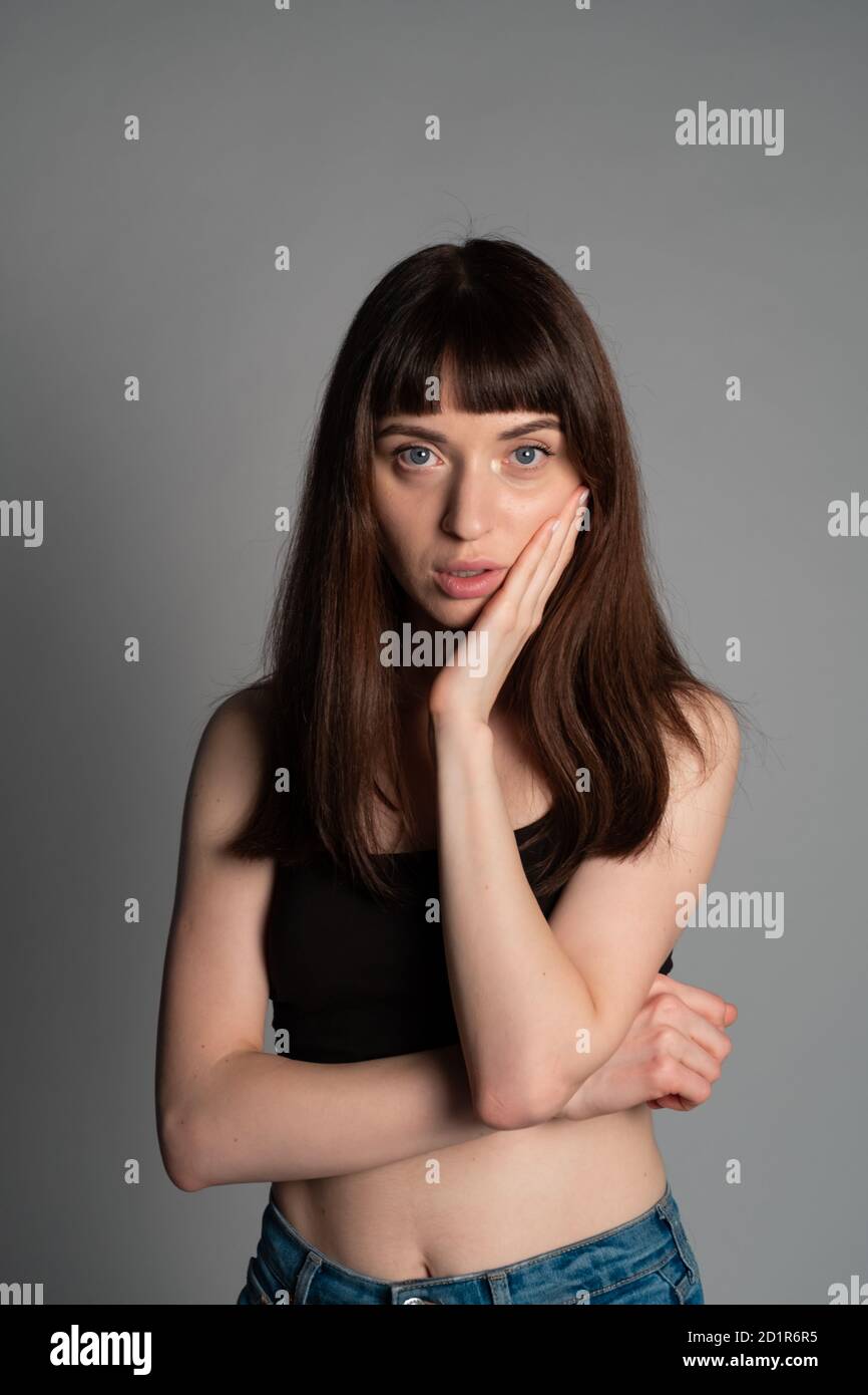 Portrait studio d'une jolie femme brunette, main sur la joue, regardant l'appareil photo, debout sur un fond gris Uni Banque D'Images