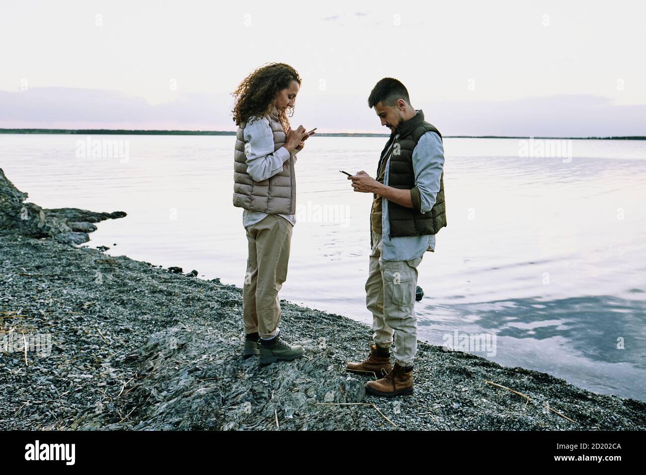 Jeune couple concentré debout sur la plage de galets et utilisant des gadgets lors de la vérification de la connexion Banque D'Images