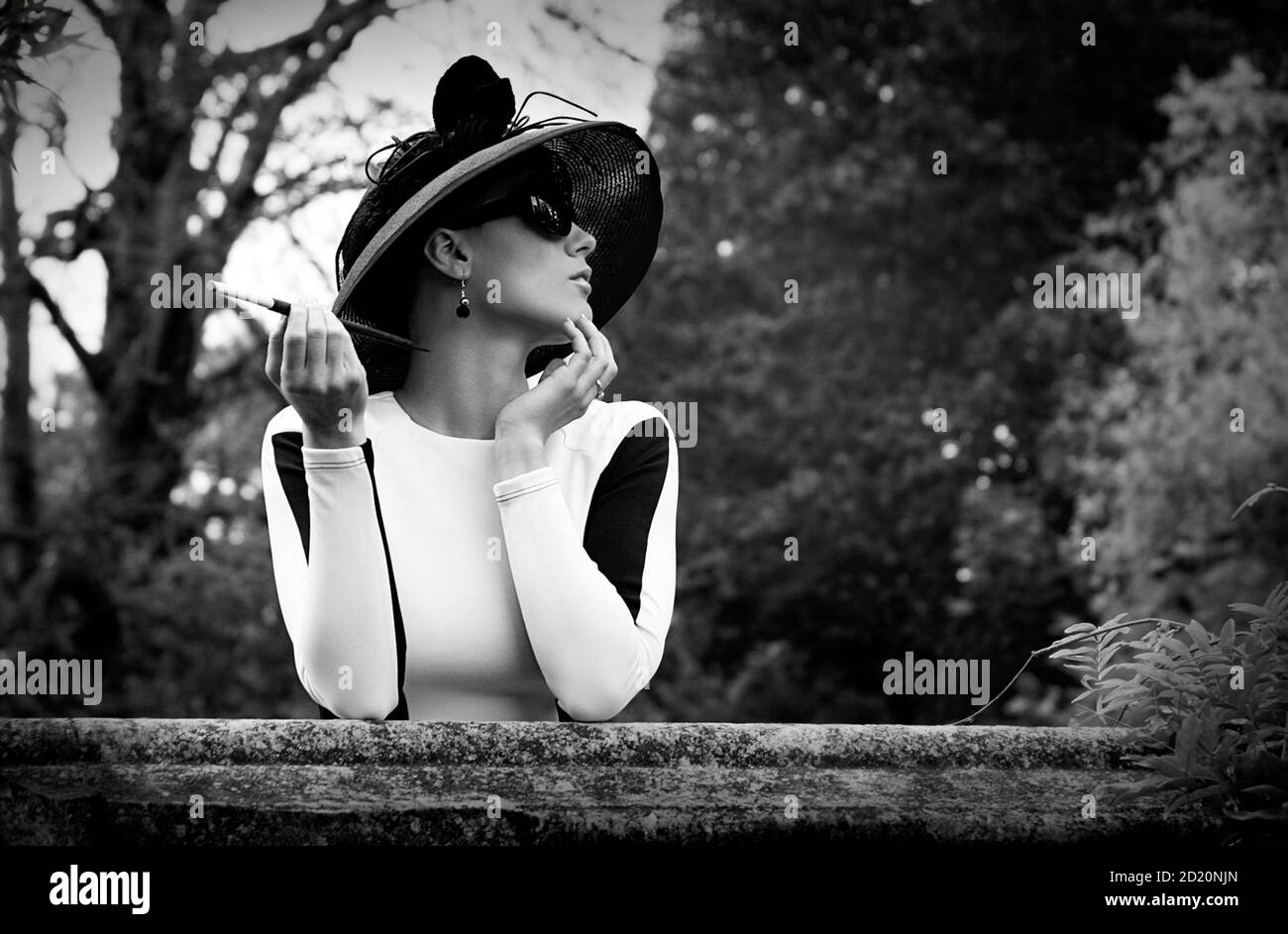 Jeune femme caucasienne dans les années 1950 Audrey Hepburn style, robe noir  et blanc bodycon, chapeau noir à large bord, lunettes de soleil noires et  un porte-cigarette Photo Stock - Alamy