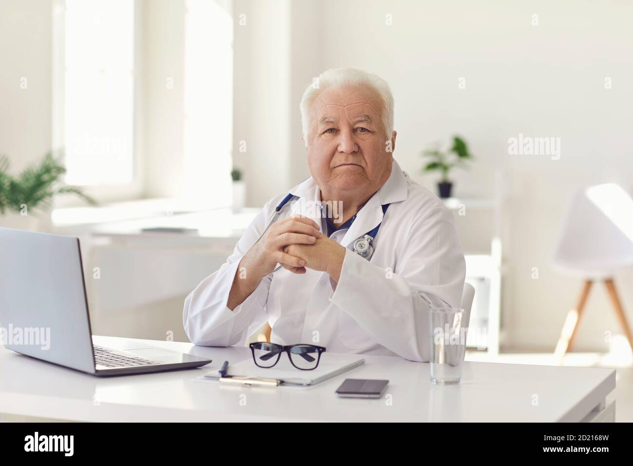 Médecin expérimenté sérieux assis à un bureau avec un ordinateur portable à l'hôpital bureau regardant la caméra Banque D'Images