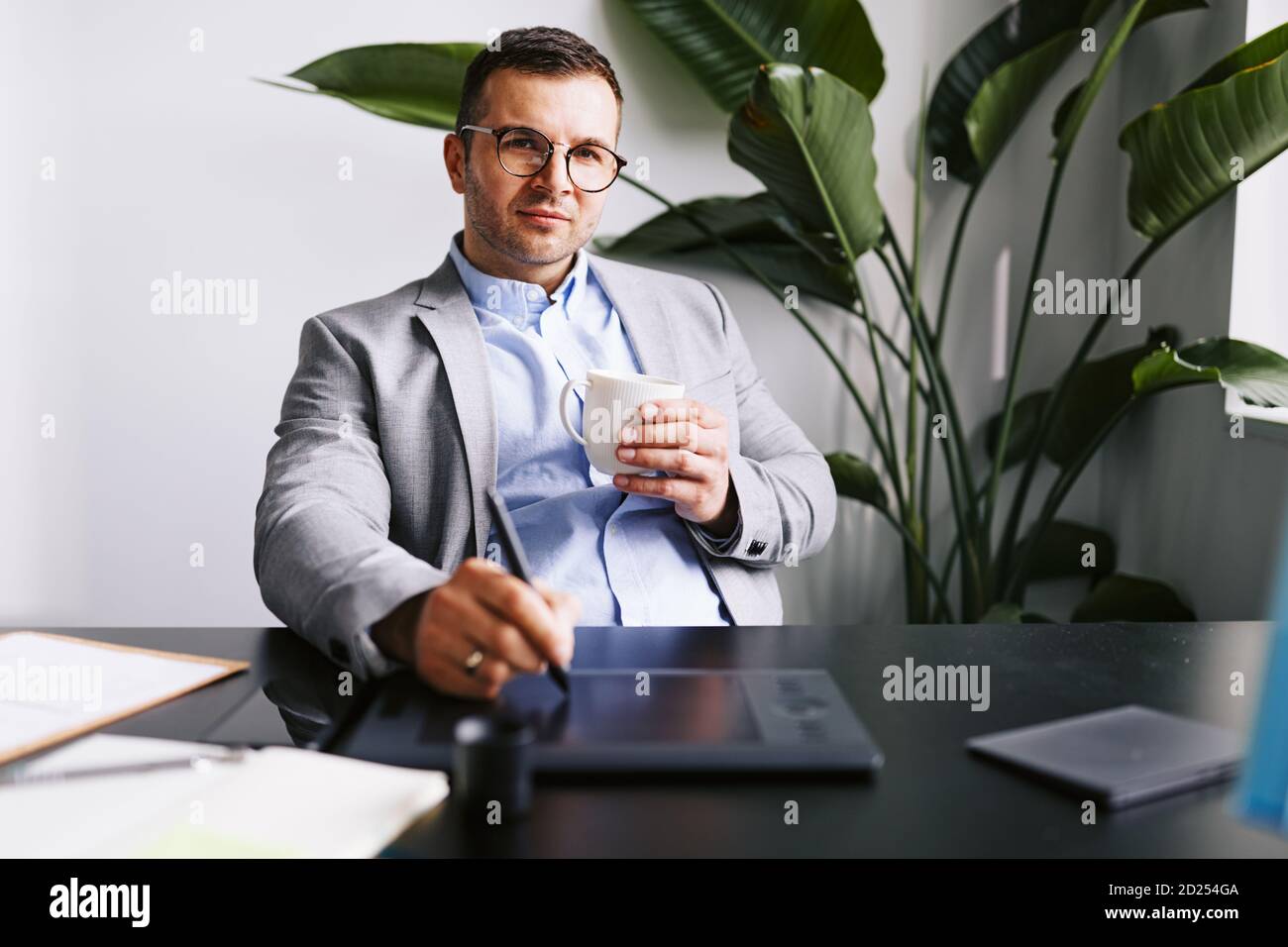 Homme d'affaires assis à son bureau buvant du café et travaillant sur un ordinateur utilisant une tablette graphique Banque D'Images