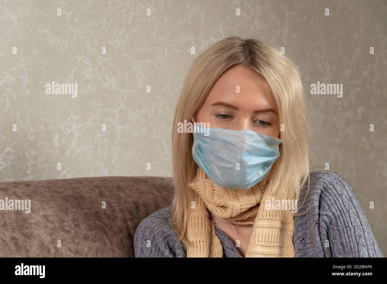 jeune femme blonde dans un masque médical avec un foulard autour de son cou est triste sur le canapé avec les yeux en bas, espace de copie Banque D'Images