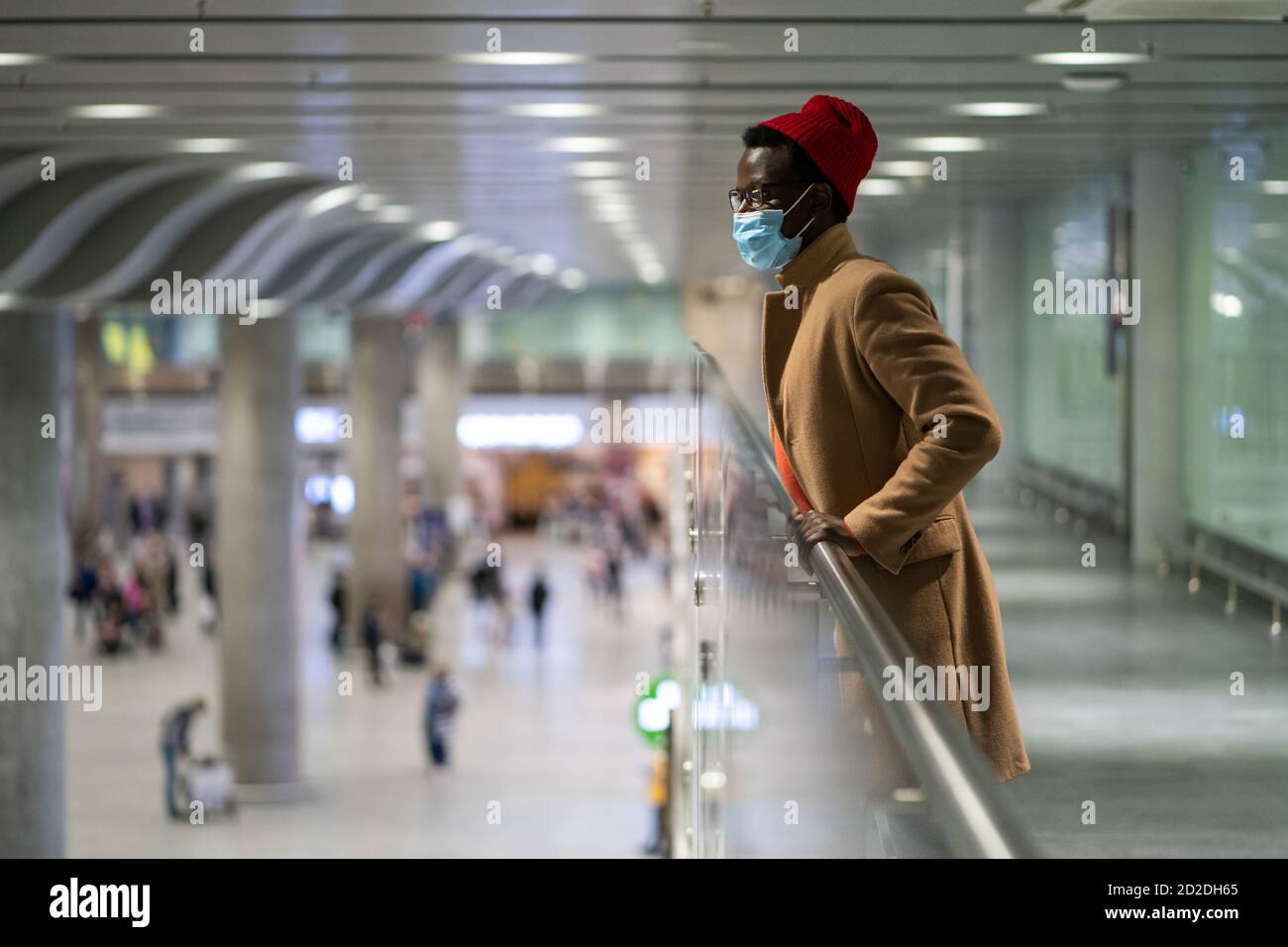 Vue latérale de l'élégant Noir voyageur homme millénaire porter masque de protection du visage, debout dans le hall de l'aéroport, regardant vers le bas, arrière-plan flou. Covid-19, Banque D'Images