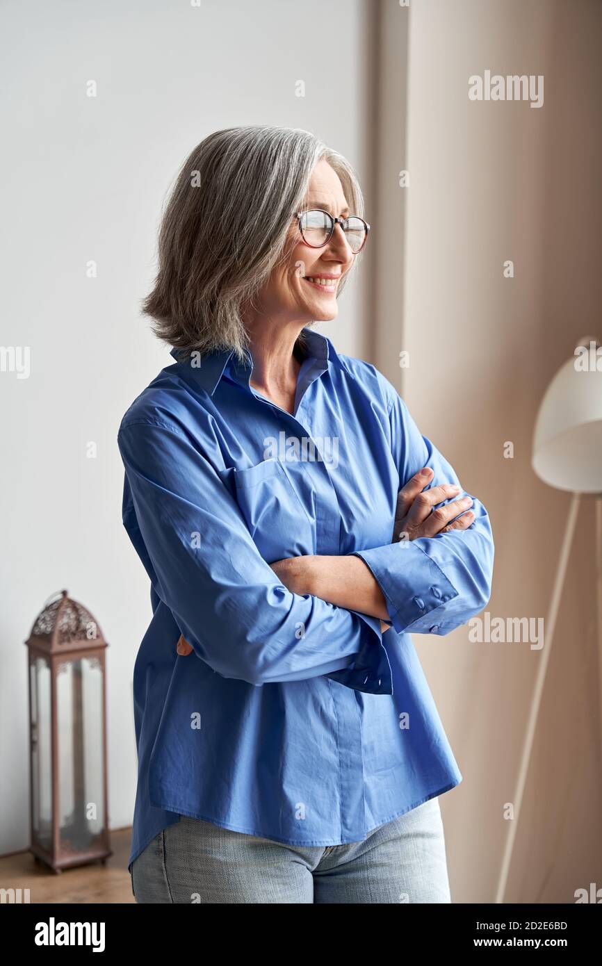 Souriante confiante femme mûre debout bras croisés à l'intérieur regardant la fenêtre. Banque D'Images