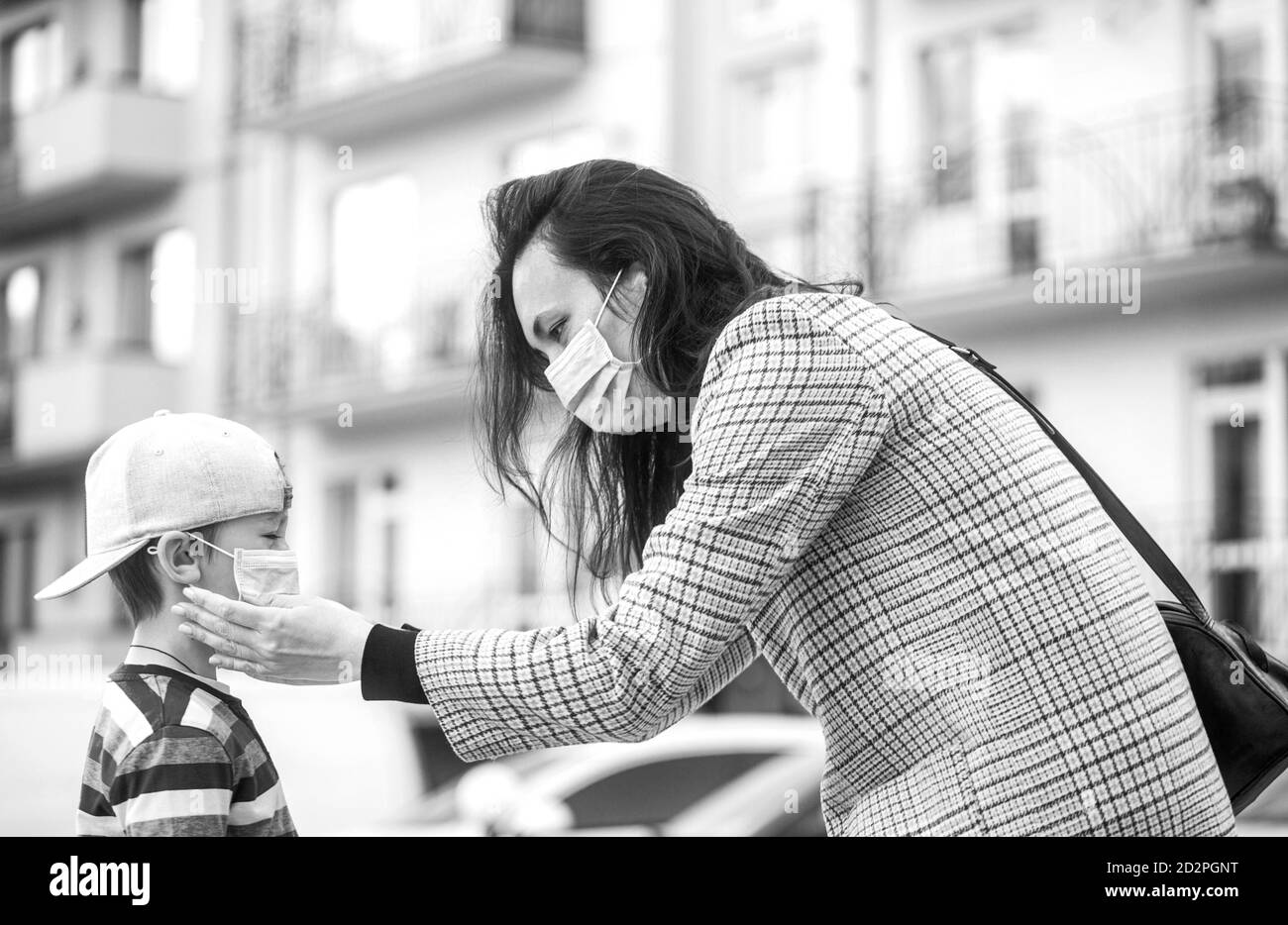 Mère met un masque de sécurité sur le visage de son fils. L'écolier est prêt à aller à l'école. Joli garçon avec un sac à dos à l'extérieur. Noir et blanc Banque D'Images