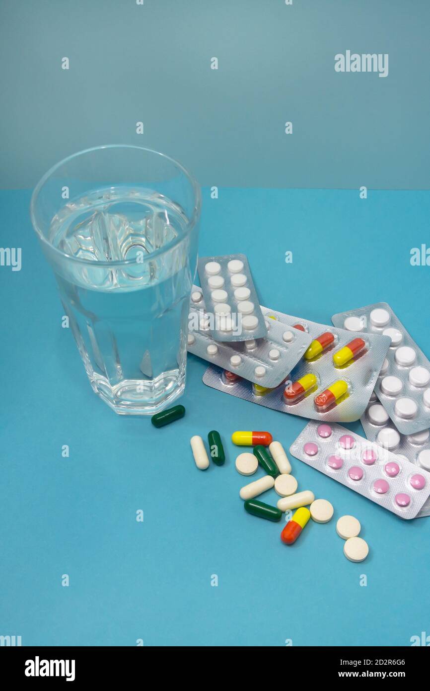 Beaucoup de pilules différentes de drogues sont dispersées et dans les cloques sur un fond bleu. Un verre d'eau propre pour les médicaments. Le concept de traitement Banque D'Images