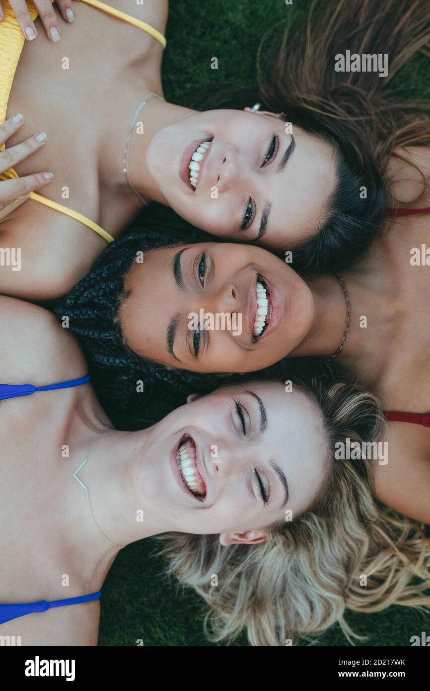 Vue de dessus de la diversité enchantée des femmes amis allongé face à faites face à une pelouse verte et regardez l'appareil photo tout en appréciant week-end ensemble Banque D'Images