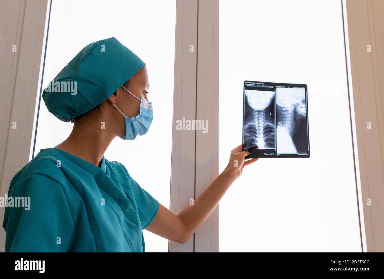 Vue latérale d'une jeune femme médecin spécialiste en blouse chirurgicale Et masque de protection examinant l'image radiographique du cou cervical colonne vertébrale du patient à l'hôpital Banque D'Images