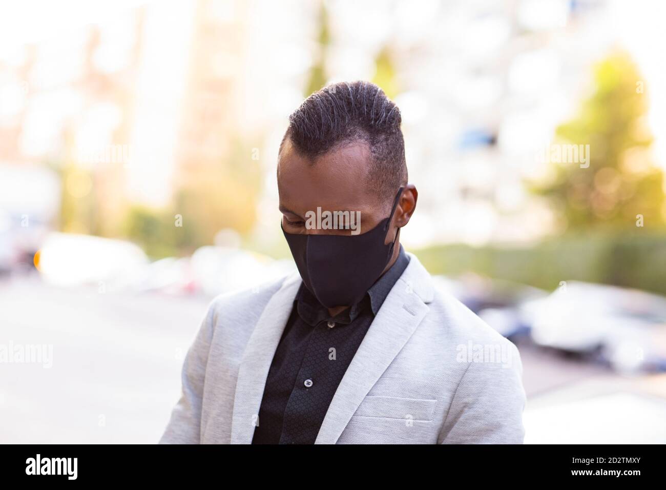 Un homme d'affaires afro-américain sérieux en costume formel et masque de protection debout dans la rue pendant la pandémie du coronavirus et en regardant vers le bas Banque D'Images