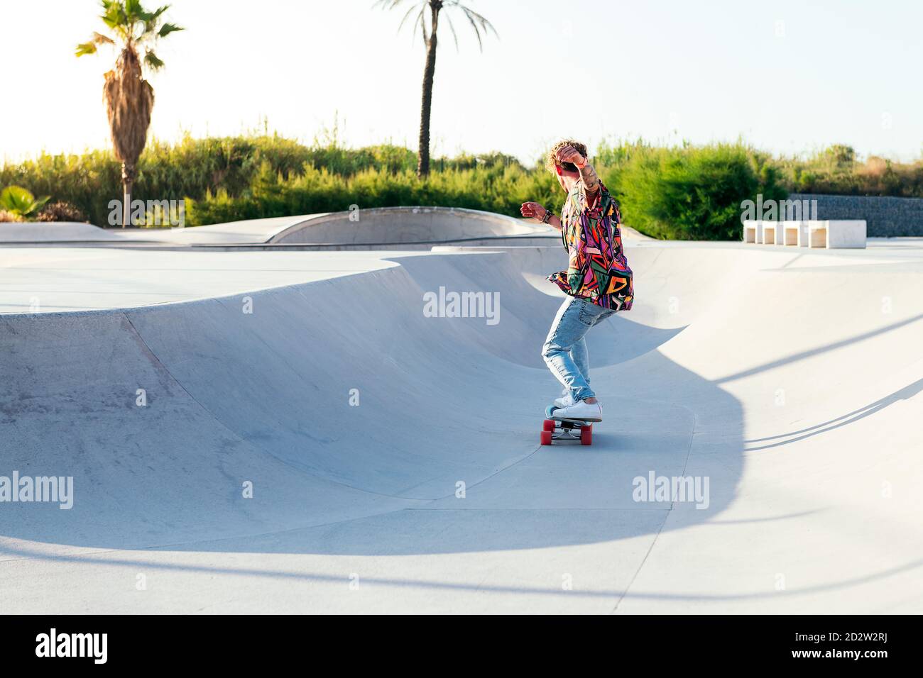 Vue arrière de l'anonyme funky jeune homme skateboarder dans le mode chemise et jean colorés en train de jouer sur une rampe en béton compétences de pratique dans le parc de skate Banque D'Images