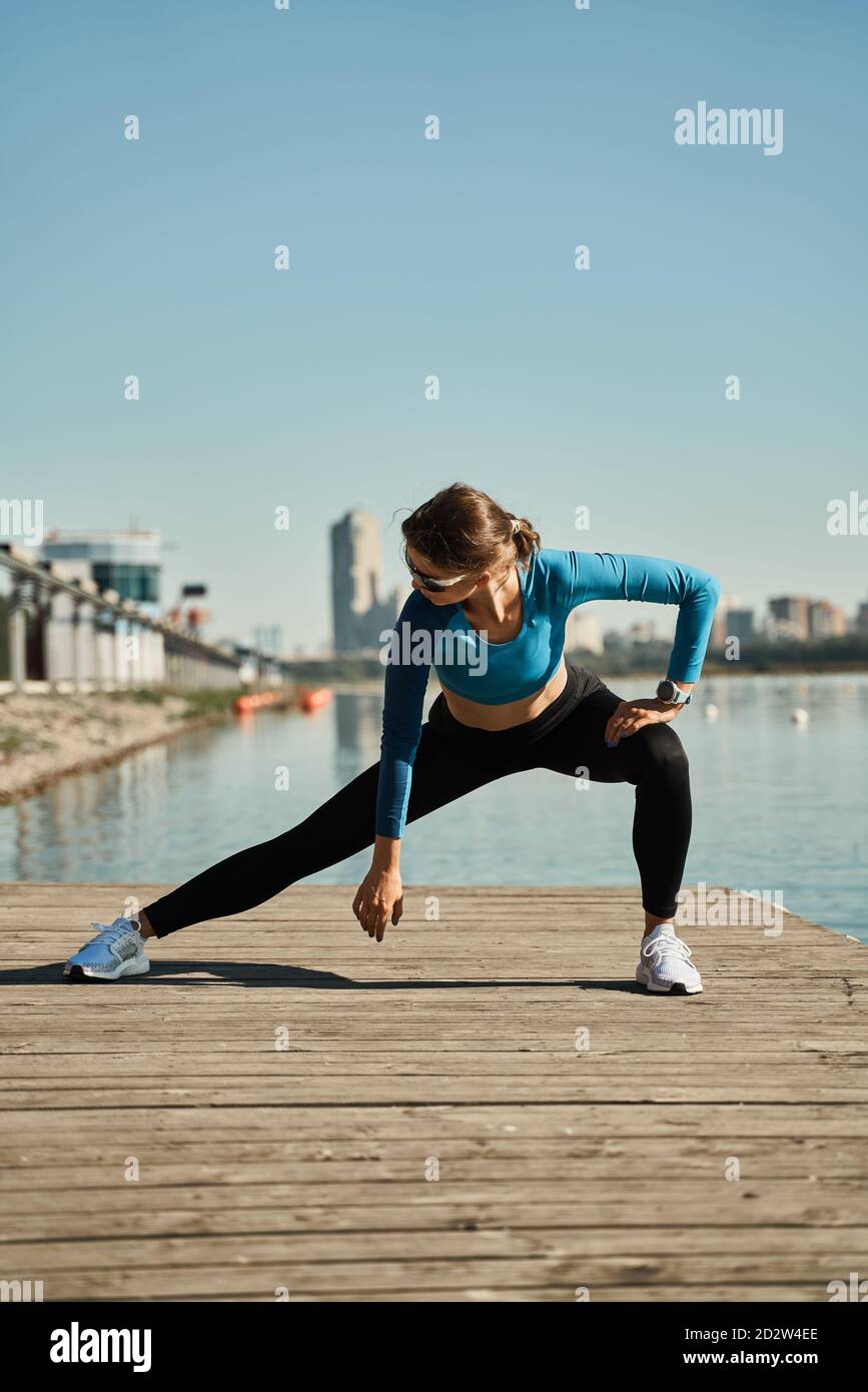 Active Slim femelle dans une tenue sportive debout sur le tapis et faire de l'exercice d'étirement pour les jambes tout en s'exerçant sur une jetée en bois près de la rivière Banque D'Images