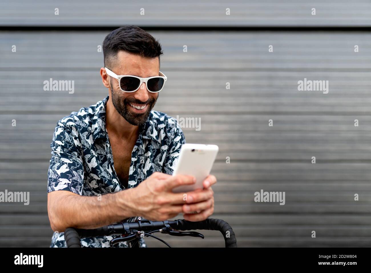 Ravi homme dans une tenue élégante penchée sur un guidon de vélo et lire des messages sur les réseaux sociaux via un téléphone mobile dans ville Banque D'Images