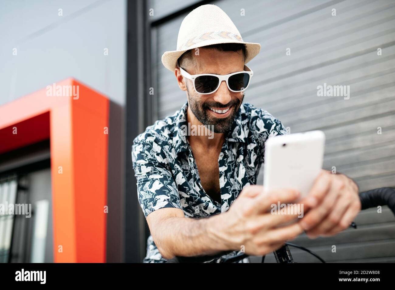 Ravi homme dans une tenue élégante penchée sur un guidon de vélo et lire des messages sur les réseaux sociaux via un téléphone mobile dans ville Banque D'Images
