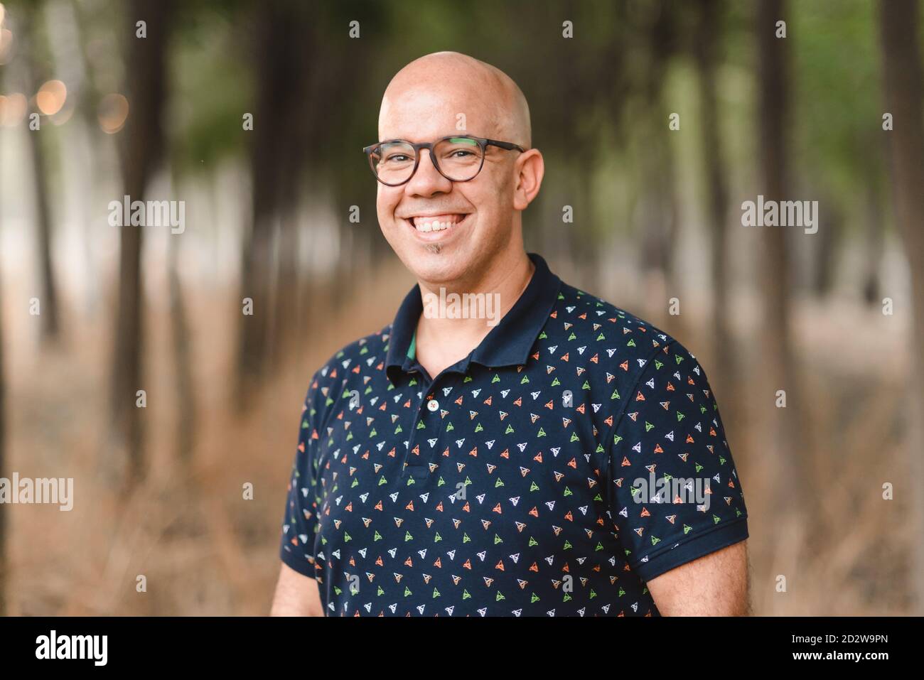 Homme beau positif avec un sourire en dents de souris et dans des lunettes debout dans une allée avec des arbres et regardant la caméra Banque D'Images