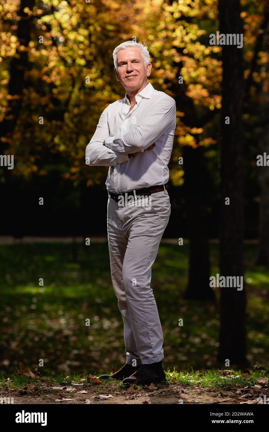 Homme mûr et calme avec une tenue élégante debout et les bras croisés dans le jardin de la ville avec des arbres jaunes et regardant la caméra Banque D'Images