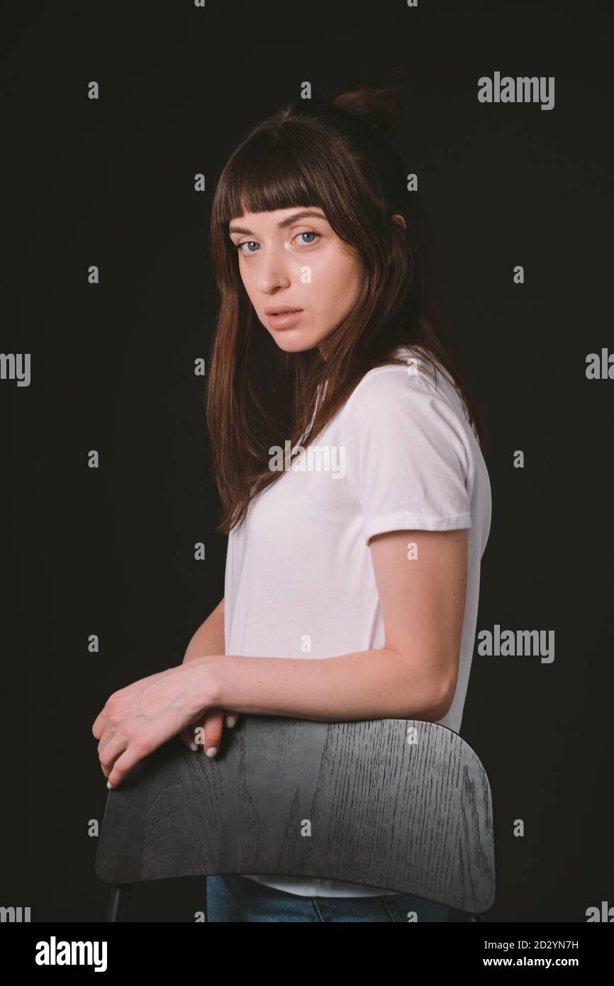 Portrait studio d'une jolie femme brune dans un t-shirt blanc, debout derrière la chaise, sur un fond noir Uni, regardant l'appareil photo Banque D'Images