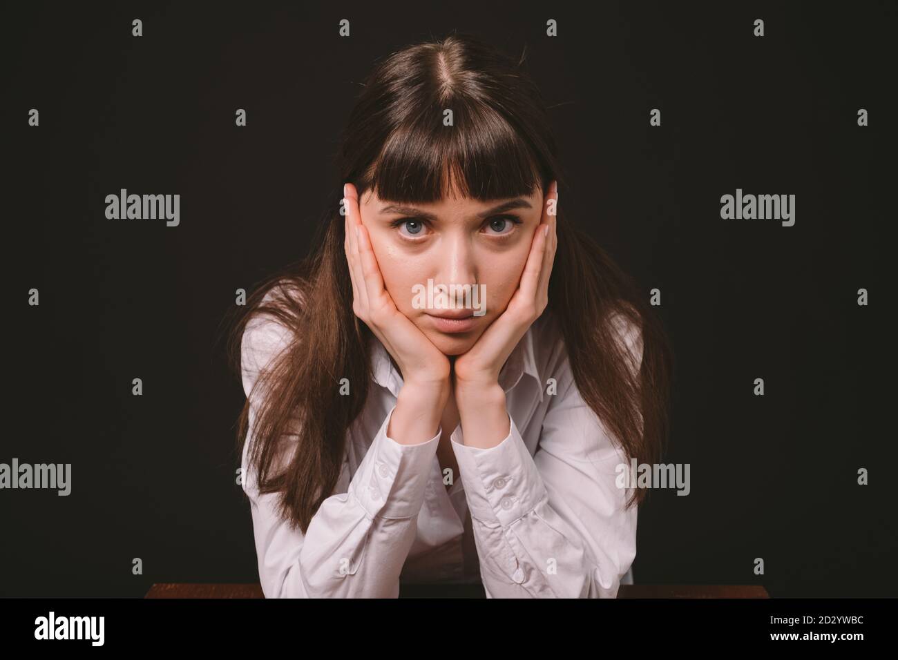 Portrait studio d'une jolie femme brune dans une chemise blanche, assise à la table, sur un fond noir Uni, regardant sérieusement l'appareil photo Banque D'Images