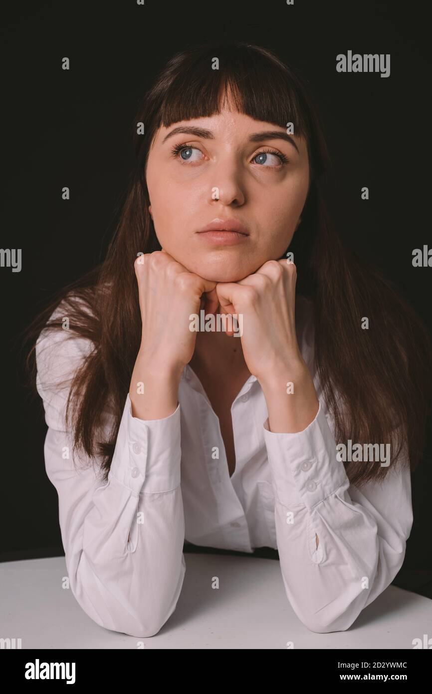 Studio portrait d'une jolie femme brune dans une chemise blanche, assise à la table, sur un fond noir Uni, regardant sur le côté Banque D'Images