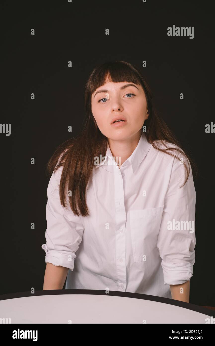 Studio portrait d'une jolie femme brune dans une chemise blanche à manches enroulées, assis à la table, sur un fond noir Uni, sérieusement Banque D'Images