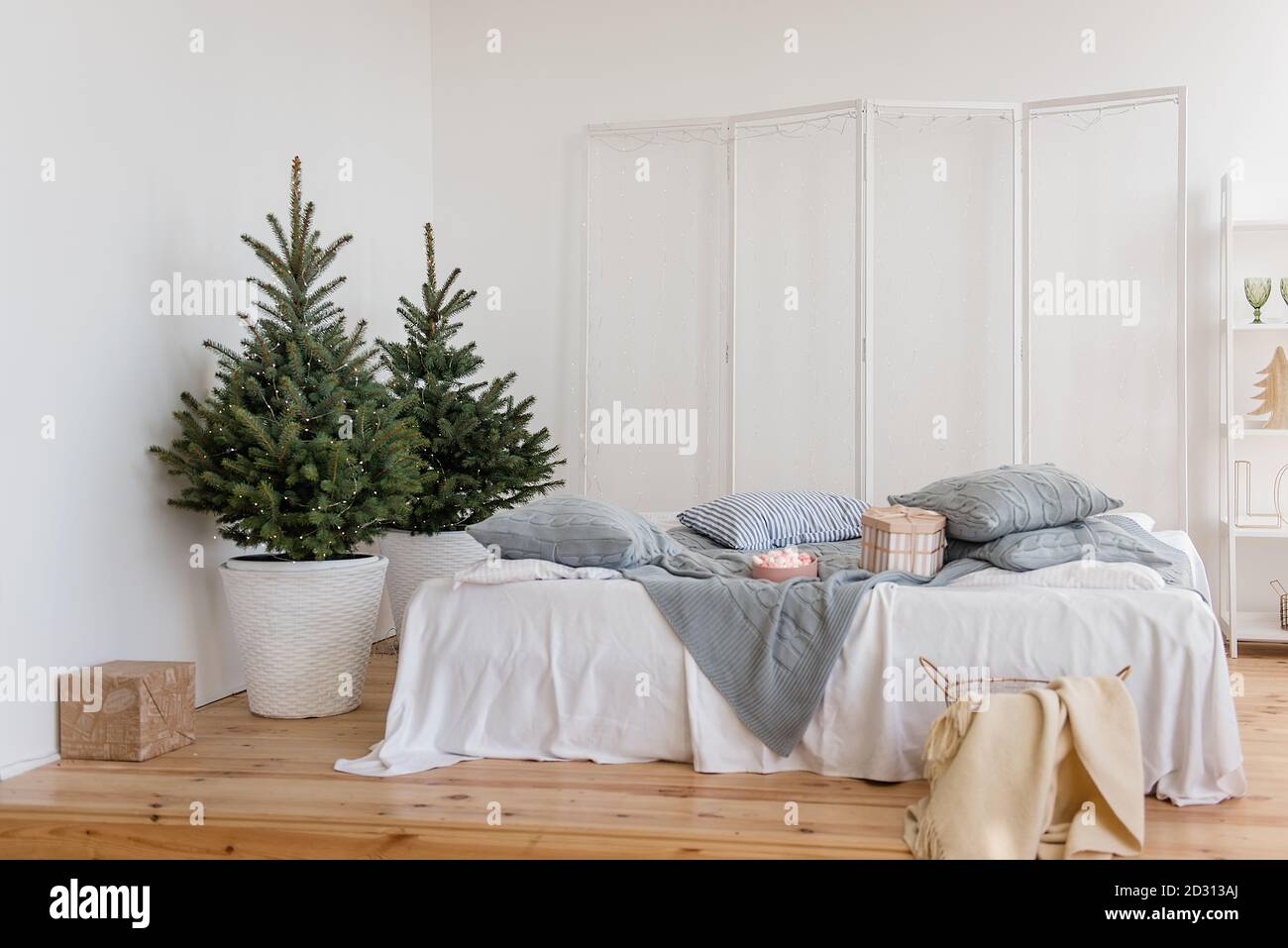 Chambre blanche élégante, minimalisme, style scandinave. Draps blancs, tissu écossais gris tricoté, oreillers. Parquet. Il y a des arbres de Noël avec guirlande Banque D'Images