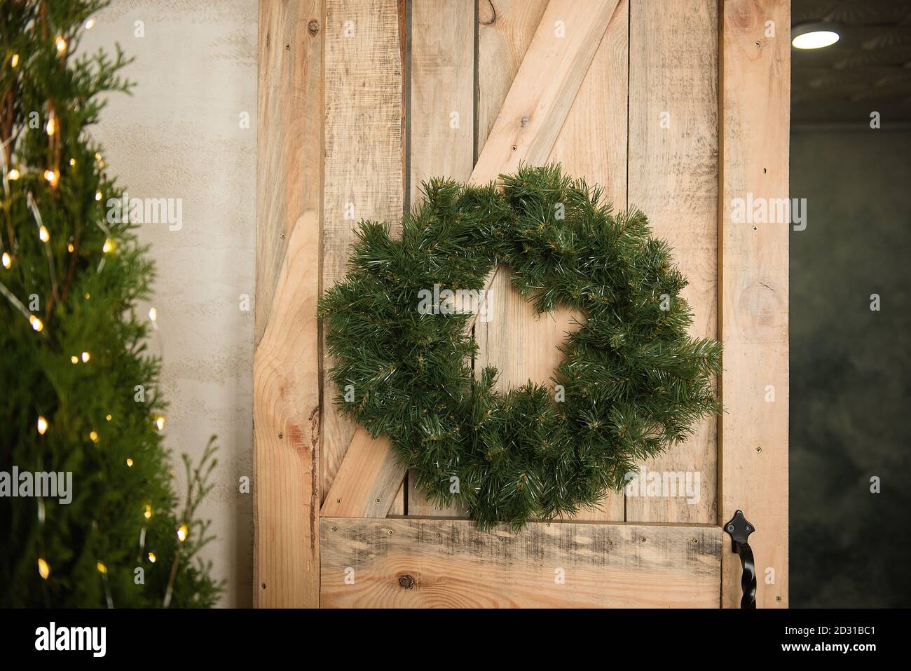 Une couronne de Noël verte en aiguilles de pin est suspendue sur la porte écologique en bois. Décoration maison festive, décorée avec une guirlande lumineuse. Texturé Banque D'Images