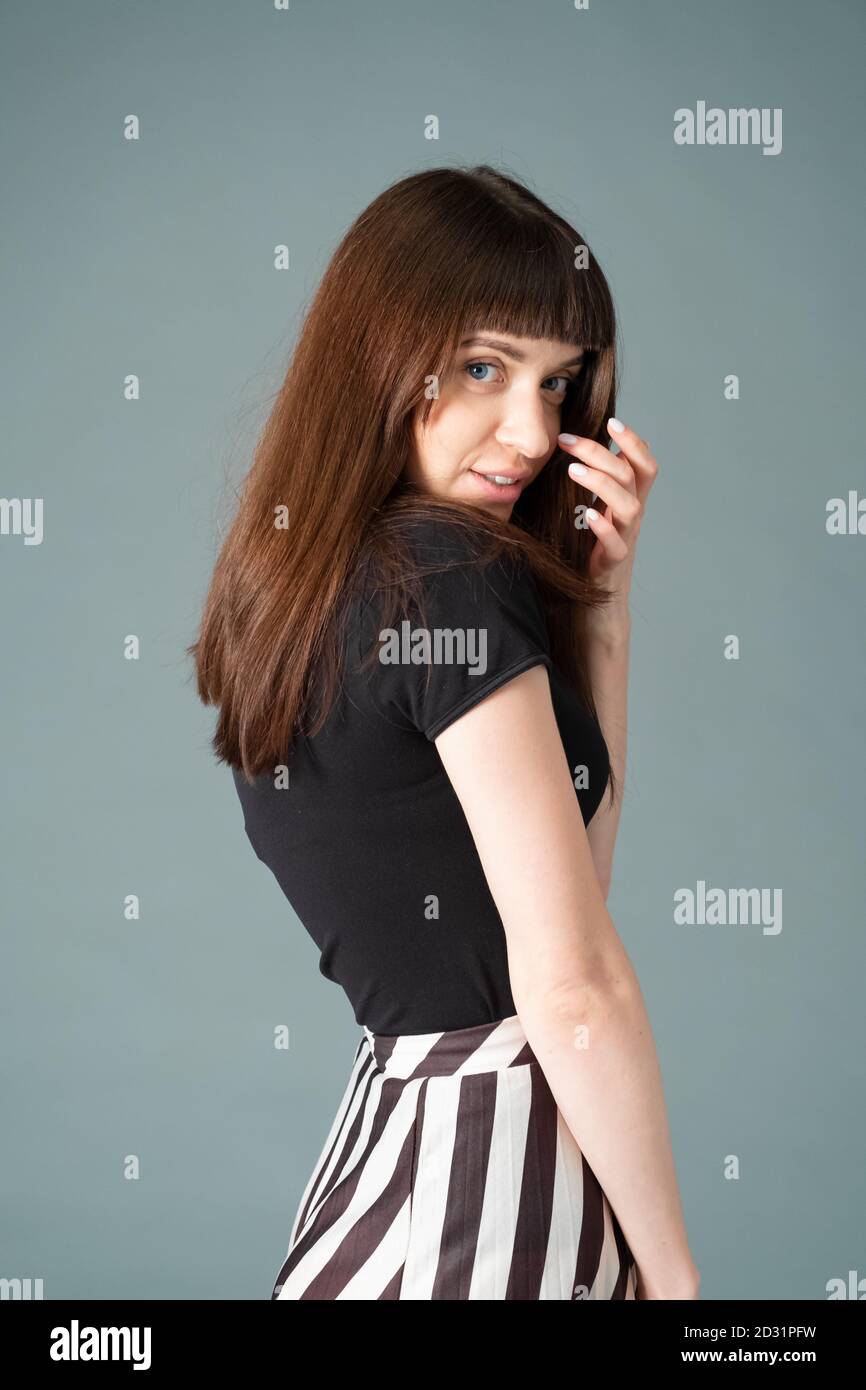 Portrait studio d'une jolie femme brune dans un t-shirt noir et une jupe rayée, debout à demi-coups, sur un fond gris Uni, regardant le Banque D'Images